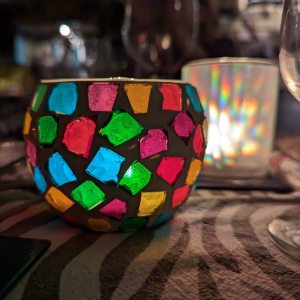 Picture of coloured glass table decoration with candle inside and glasses plus another night light in the background